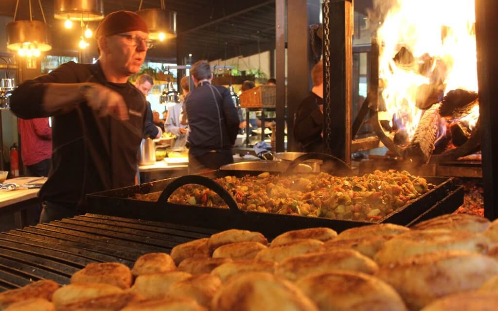 Ein Koch wendet geschmortes Gemüse in einer großen rechteckigen Grillpfanne. Im Hintergrund sind man Gäste am Buffet und ein offenes Holzfeuer, das den Grill anheizt